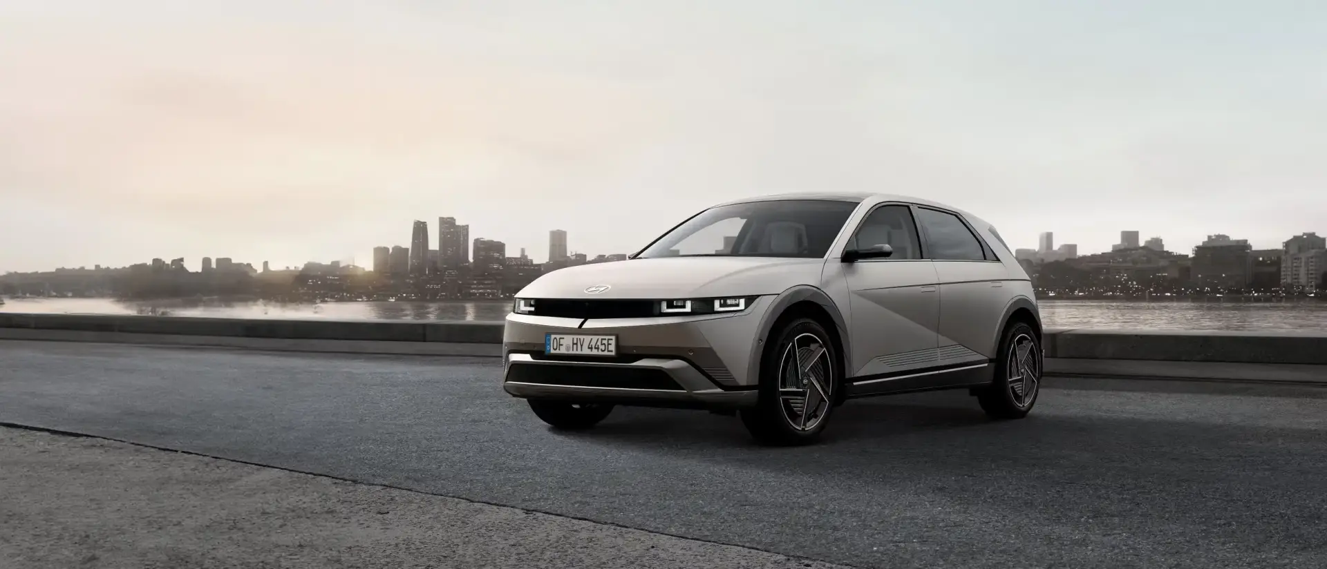 Hyundai Ioniq 5 electric SUV parked by a waterfront road with a city skyline in the background during sunset, highlighting its futuristic design and eco-friendly focus.